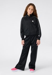 Jong meisje met lang, krullend haar, gekleed in een zwarte trainingsoverall met witte piping en roze sneakers, staand tegen een effen achtergrond.