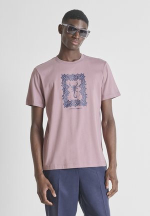 Joven con gafas de sol grises, camiseta color malva con gráfico de escorpión azul oscuro y pantalones azul marino sobre un fondo claro y simple.