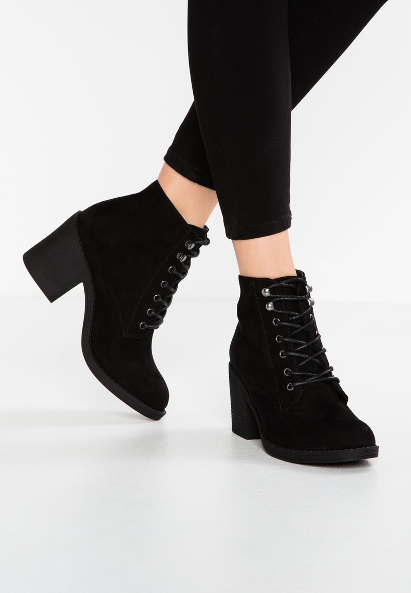 Bottines en daim noir avec des talons carrés et des lacets, portées avec un pantalon noir ajusté sur un sol blanc.