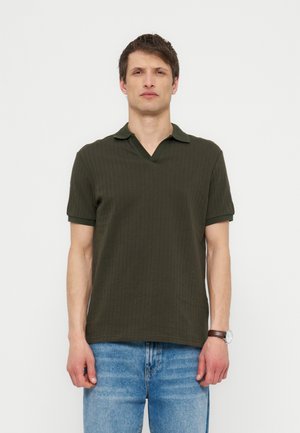 Muž stojící čelem vpřed, oblečený v tmavě zeleném žebrovaném polo tričku s krátkým rukávem, modrých džínách a náramkových hodinkách na levém zápěstí.