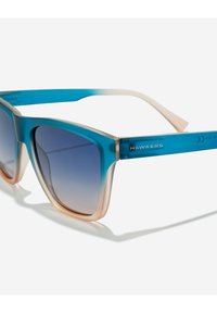 Modre in bež gradientne pravokotne sončna očala z modrimi zatemnjenimi lečami in blagovno znamko "Hawkers" na ročki.