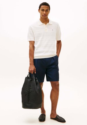 Hombre que lleva un polo blanco de manga corta y pantalones cortos azul marino, sosteniendo una bolsa tote negra en una mano y con la otra mano en el bolsillo.