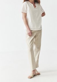 Blouse blanche à manches courtes avec décolleté en V et motif brodé, accompagnée d'un pantalon en lin beige clair et de sandales simples couleur tan.