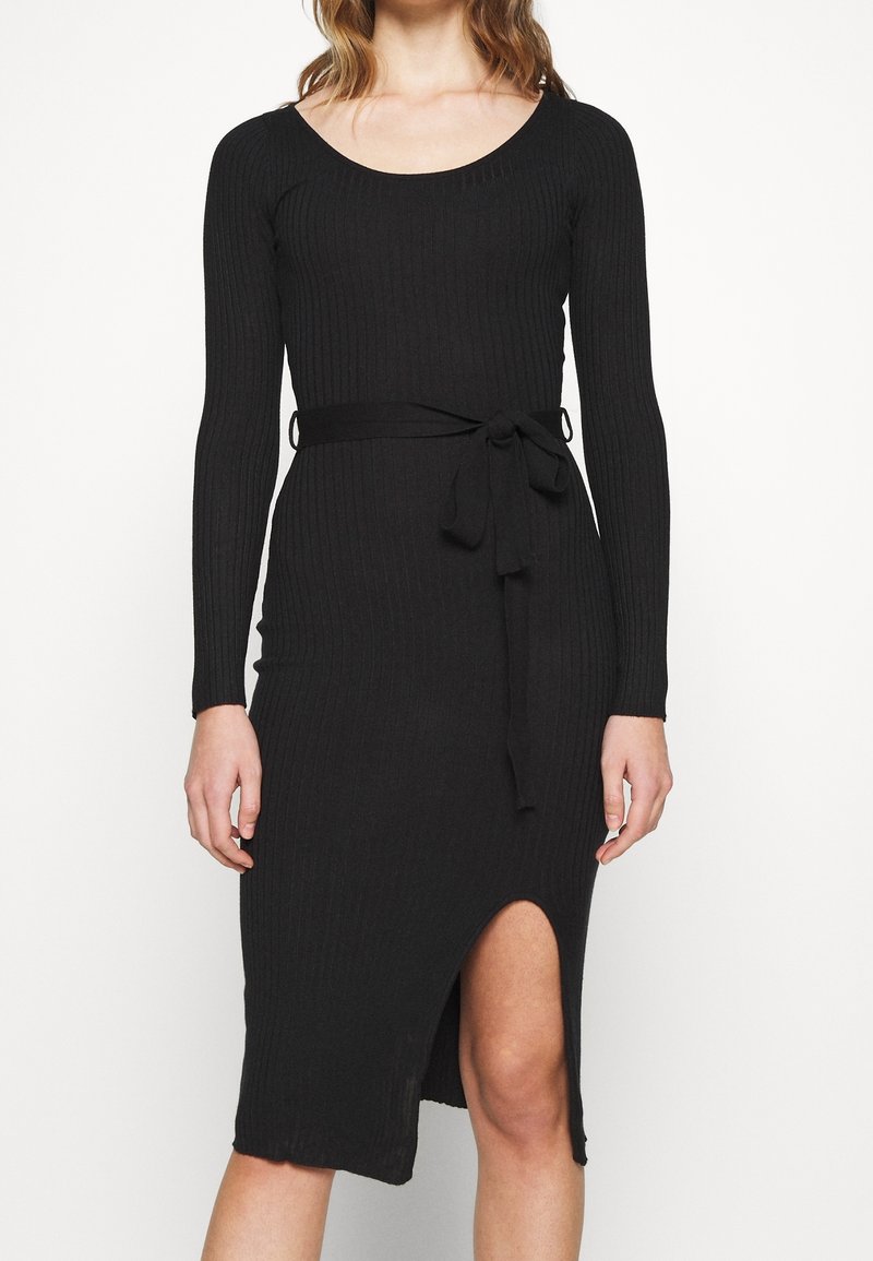 Femme portant une robe noire en maille côtelée avec manches longues, un décolleté en arrondi, une ceinture nouée à la taille et une fente devant au-dessus du genou.