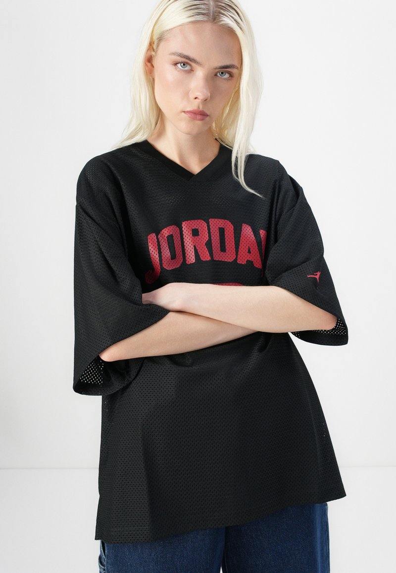 Maillot en mesh noir avec des manches larges, arborant "JORDAN" en lettres rouges. Design en V avec un petit logo sur la manche. Tissu texturé.
