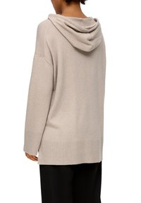 Osoba ubrana w beżowy, dziergany sweter z kapturem, z długimi rękawami i ściągaczami przy mankietach, widziana od tyłu, w połączeniu z czarnymi spodniami.