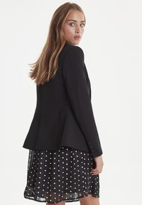 Černý blazer se strukturovanými rameny a rozšířeným zadním dílem, nošený přes průhlednou černou sukni s bílými puntíky. Má zapínací manžety.