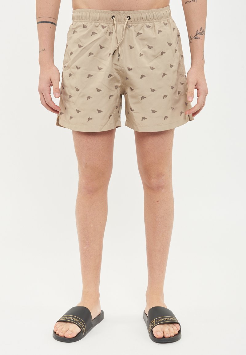 Costumi da bagno beige con piccoli motivi geometrici marroni, vita elastica con cordino e tasche laterali. Indossati con sandali neri a ciabatta.