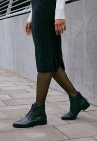 Botines de piel negra con diseño de punta afilada, detalle de cordones y cremallera lateral. Llevados con un vestido negro de canalé y medias transparentes.