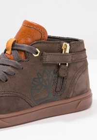 Zapato alto de ante marrón con un acento de cuero naranja, cordones grises y detalles en cremallera dorada. Presenta un logo de árbol en el lateral.