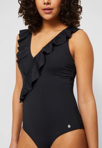 Maillot de bain noir avec un décolleté en V profond, une épaule avec des volants, et une finition lisse et élégante. Petit accent métallique à la taille.