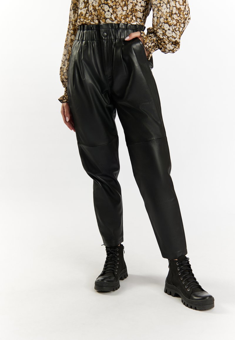 Pantaloni high-waisted in pelle nera con vestibilità rilassata, caratterizzati da pieghe e vita elasticizzata, abbinati a stivali da combattimento neri con lacci.