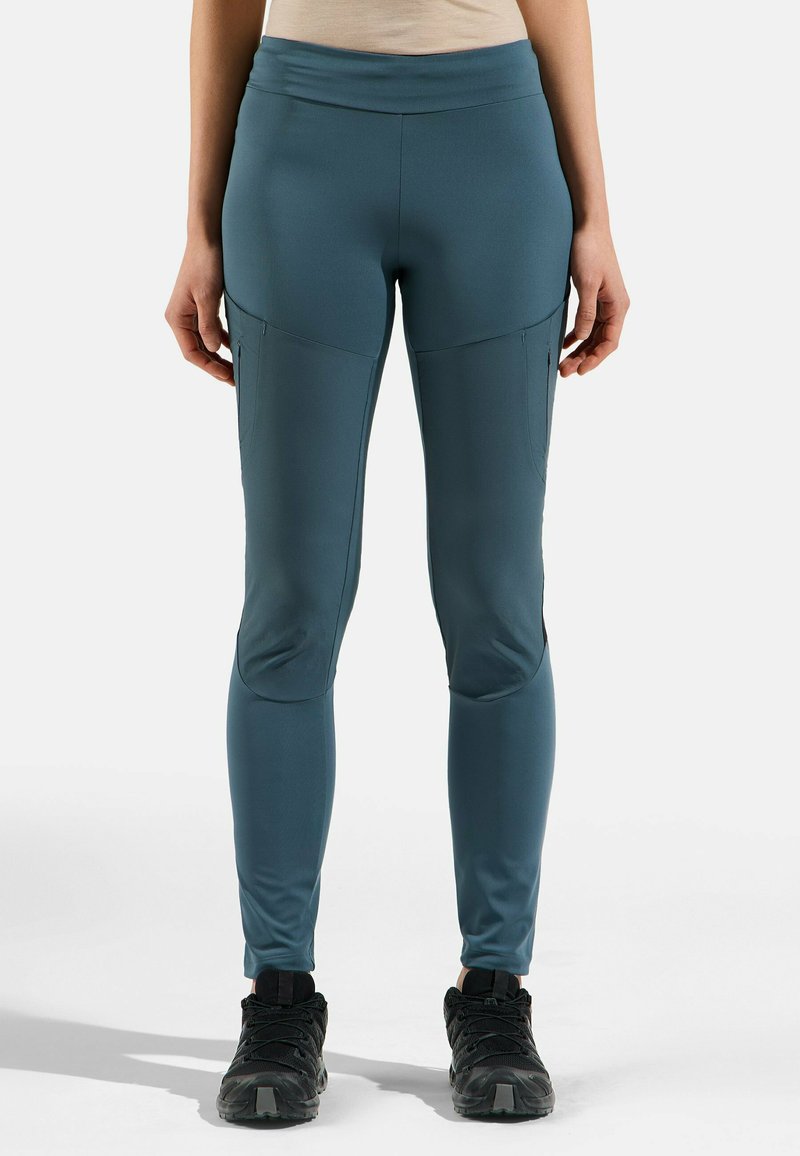 Leggings de sport en tissu teal, design près du corps avec rembourrage aux genoux, poches latérales zippées et taille élastique large. Chaussures noires.