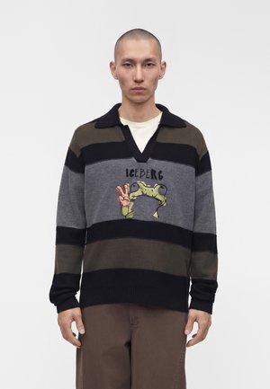 Junger Mann trägt einen schwarz-grau-oliv gestreiften Pullover mit einer Froschgrafik, die ein Friedenszeichen zeigt, und dem Schriftzug "ICEBERG" auf der Brust.