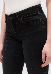Fekete farmernadrág, testhez simuló szabással, apró fém szegecsekkel díszítve a szöveten, és klasszikus gombos záródással deréknál.