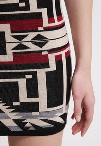 Gros plan de la main d’une personne à côté d’une jupe avec un motif géométrique en noir, rouge, beige et gris.