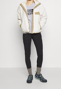 Fleece bunda v krémové barvě s béžovými akcenty, šedé tričko, černé kalhoty s kapsami a modro-zelené sportovní boty s texturovanou podrážkou.
