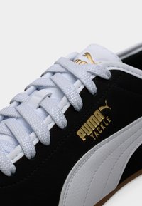 Κοντινό πλάνο μαύρου και λευκού αθλητικού παπουτσιού Puma με χρυσό λογότυπο "Puma Tackle" και λευκά, υφασμένα κορδόνια.