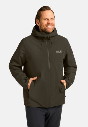Mann lächelt und trägt eine dunkelgrüne Jack Wolfskin Kapuzenjacke mit den Händen in den vorderen Taschen vor einem einfarbigen Hintergrund.