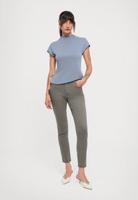 Kvinne med mørkt hår og tatoveringer iført en blå kortermet topp, grå slim-fit bukser og hvite lave hæler, stående med en hånd i lommen.