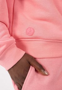 Lichtroze sweatshirt met een zachte textuur, voorzien van een rond logopatch met het label "JOTT." Inclusief een voorzak en details met elastische manchetten.