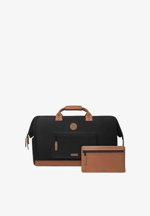 Sac de voyage noir et marron avec poche avant et pochette marron zippée assortie reposant devant sur fond blanc.