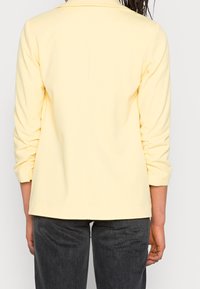 Blazer amarillo con un diseño estructurado, mangas largas y una textura suave. La parte de atrás es lisa, sin detalles visibles. Combinado con pantalones oscuros.