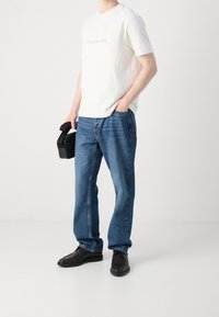 Bílé bavlněné tričko s logem Calvin Klein Jeans, v kombinaci s modrými džíny. Černé boty. Muž drží černou tašku s držadlem.