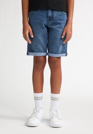 Pantaloni scurți din denim, de culoare albastru mediu, cu tiv întors, combinați cu adidași albi și șosete cu dungi. Design simplu, croială lejeră și lungime până la mijlocul coapsei.