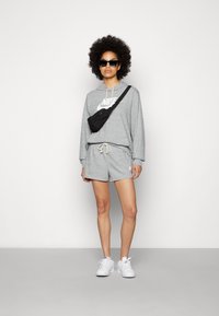 Felpa grigia con logo Nike bianco, pantaloni corti grigi abbinati, borsa a tracolla nera e sneakers bianche. Modello con capelli ricci e occhiali da sole.