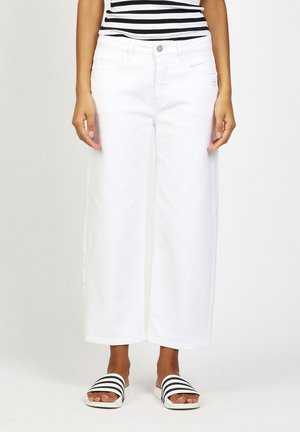 Frau trägt weiße, weit geschnittene, verkürzte Hosen, schwarz-weiß gestreiftes Oberteil und dazu passende gestreifte Sandalen, steht vor einfarbigem Hintergrund.