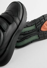 Chaussures en cuir noir avec deux bandes Velcro. Semelles en caoutchouc épaisses avec des accents verts et orange. Détails texturés sur la semelle pour une meilleure adhérence.