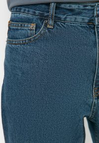 Vaqueros de mezclilla azul con superficie texturizada, con costuras naranjas, cinco bolsillos y una cintura de corte medio.