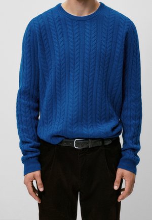 Homme portant un pull en maille torsadée bleu vif rentré dans un pantalon noir avec une ceinture noire, mains détendues le long du corps, devant un fond uni.