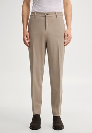 Mann trägt beige maßgeschneiderte Hosen mit Frontfalz, kombiniert mit einem weißen Hemd und dunkelbraunen Stiefeletten vor einfarbigem Hintergrund.