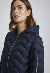 Marinblå pufferjacka med ett chevron-quiltat mönster, dragkedja fram och huva. Har sidofickor och en silverfärgad dragkedjedetalj.