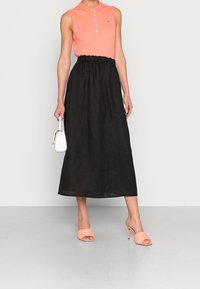 Frau trägt ein korallenfarbenes ärmelloses Poloshirt, einen schwarzen Hochglanz-Midi-Rock und korallenfarbene Absatzsandalen, während sie eine kleine weiße Handtasche hält.