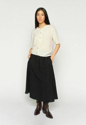 Nainen seisoo käsillä taskuissa, pukeutuneena kermaiseen, lyhythihaiseen napitettuun neulepuseroon, mustaan midihameeseen ja tummanruskeisiin nahkasaappaisiin.