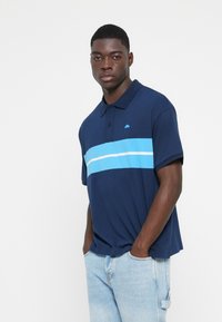 Mörkblå pikétröja med en horisontell ljusblå och vit rand över bröstet, tillverkad av bomullsblandning, med krage och korta ärmar.