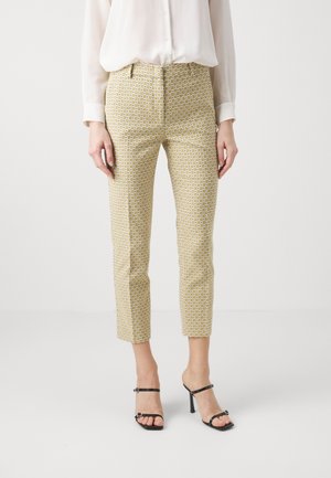 Mujer que lleva pantalones cortos beige con estampado, una blusa blanca de manga larga y sandalias negras de tacón alto, de pie frente a un fondo liso.