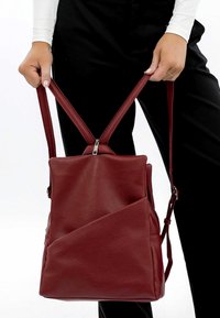 Roter Leder-Rucksack mit strukturiertem Oberflächen, asymmetrischem Design, zwei verstellbaren Tragegurten und Reißverschluss. Verfügt über eine zusätzliche Seiten Tasche.
