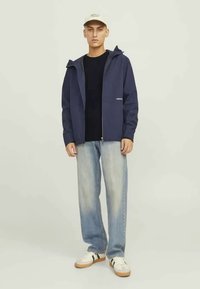 Mörkblå zip-up hoodie med en logotyp, bärs över en svart t-shirt. Ljust blå denimjeans och vita sneakers med gröna ränder. Beige keps.