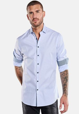 Mann mit kurzen Haaren und Tätowierungen trägt ein hellblaues Hemd mit hochgekrempelten Ärmeln und schwarze Jeans vor einfachem Hintergrund.