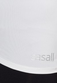 Tissu blanc avec des coutures visibles au-dessus d’un tissu noir, avec un petit logo argenté « casall » en bas à droite.