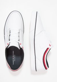 Baskets blanches présentant une texture en cuir lisse, une semelle extérieure noire, des accents rayés rouges et blancs, ainsi que des lacets sur une languette rembourrée.