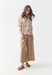 Camisa abotonada color beige claro con patrones de palmeras bordados, combinada con pantalones anchos sueltos color arena y sandalias planas marrones. Textura suave.