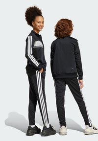 Survêtement de sport noir avec des rayures blanches ; veste à fermeture éclair à l'avant et pantalon fuselé ; texture lisse ; panneau gris réfléchissant sur la poitrine.