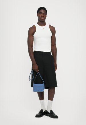 Mann in weißer ärmelloser Bluse, schwarzen knielangen Shorts, weißen Socken, schwarzen Slippern, der eine kleine blaue Handtasche vor einem weißen Hintergrund hält.