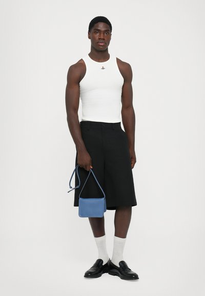 Mand iført hvid ærmeløs top, sorte knælange shorts, hvide sokker, sorte loafers, der holder en lille blå håndtaske mod en hvid baggrund.