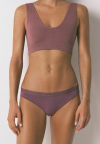 Donna che indossa un reggiseno sportivo malva e mutandine abbinate con testo "women’secret" sulla cintura, in piedi contro uno sfondo chiaro uniforme.